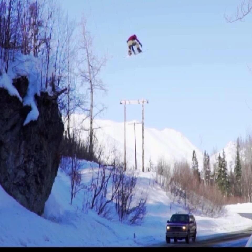 A snowboarder grabbing the board mid-air while clearing the Hatcher Pass Canyon Gap jump in Alaska, flying over a parked car below.