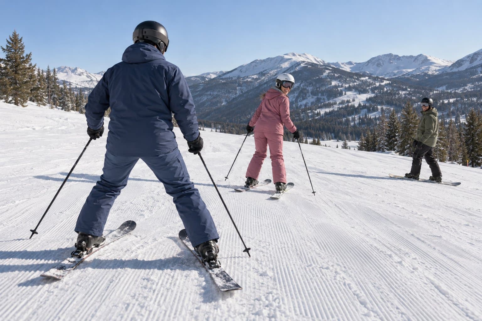 An experienced skier rides slowly in front of a newer skier in a snowplough on a gentle groomed run while a snowboarder waits below.
