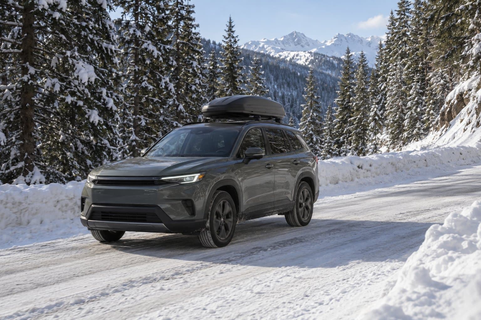 An SUV with a roof box drives along a plowed mountain road between snowbanks and evergreen trees.