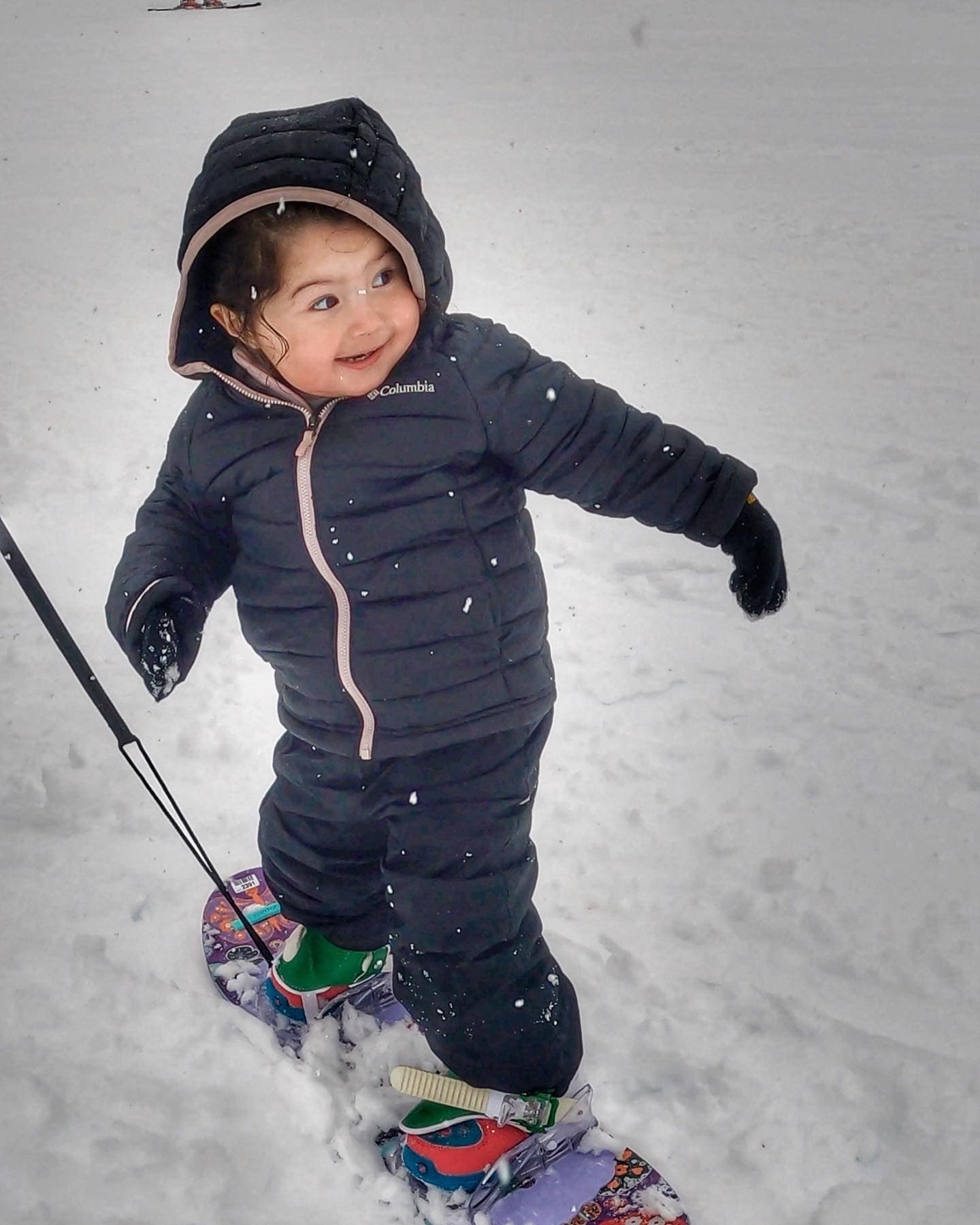 A toddler standing on a snowboard on a gentle beginner slope, learning to ride with a parent close by.