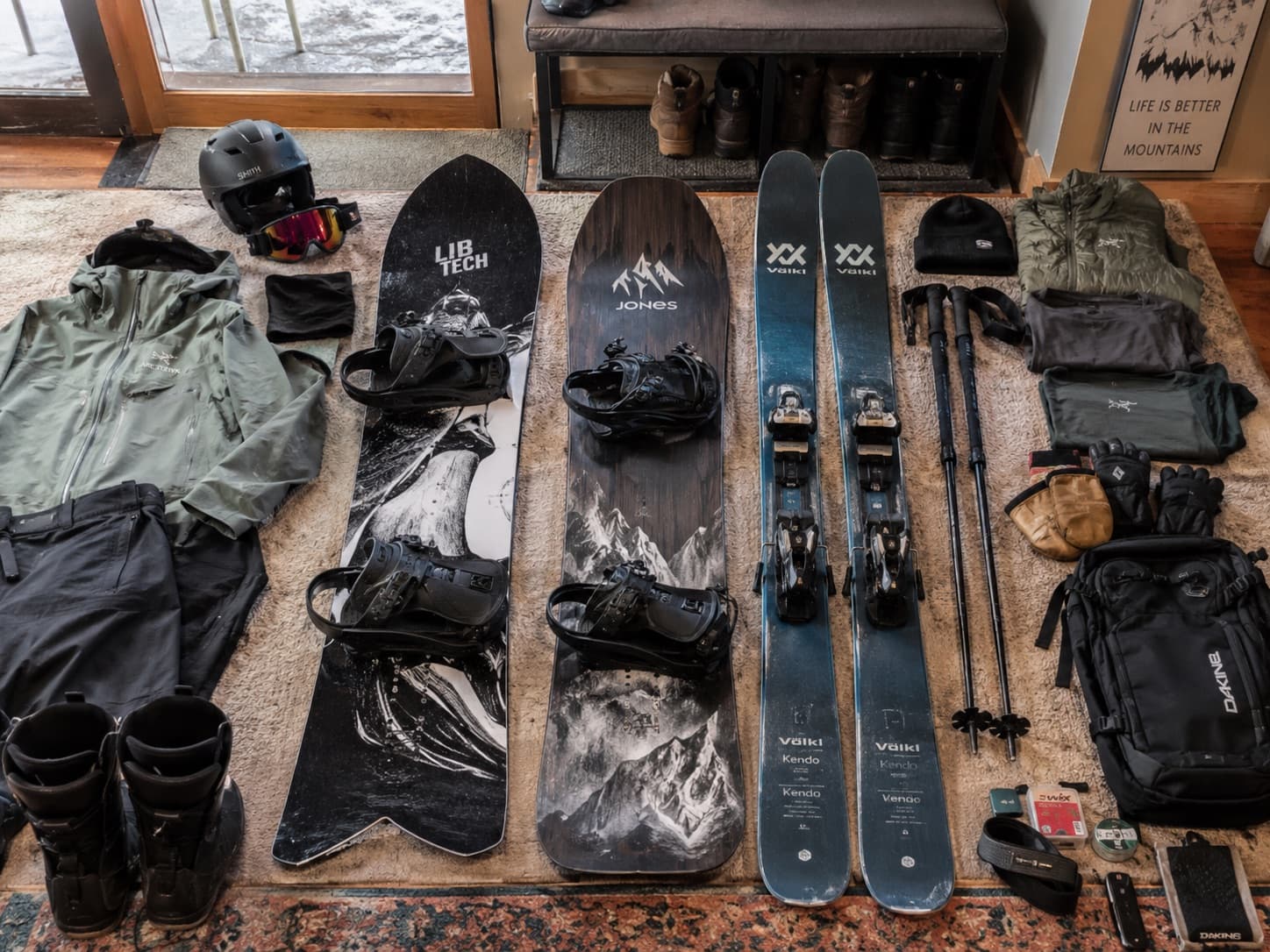 Ski and snowboard gear laid out on a rug by a snowy door before the season: two snowboards with bindings, a pair of skis with poles, a helmet and goggles, a shell jacket and pants, base layers, gloves and mittens, snowboard boots, a backpack and a wax kit.