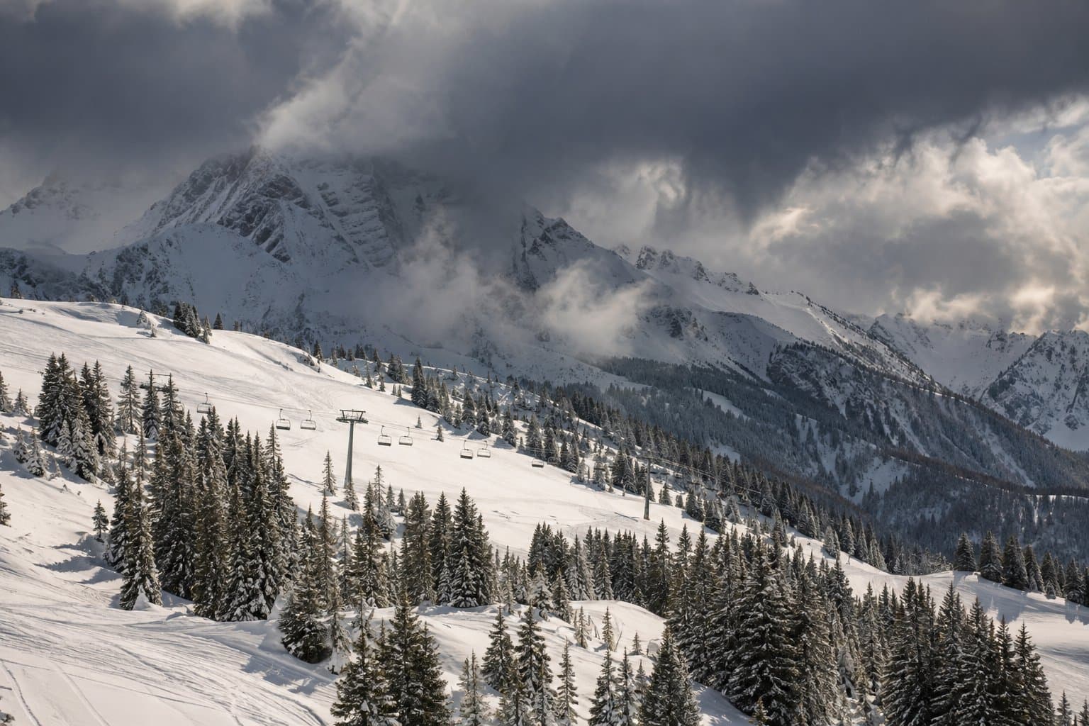 A winter storm rolls over snowy peaks above a ski area while sun still lights the lower slopes and a chairlift.