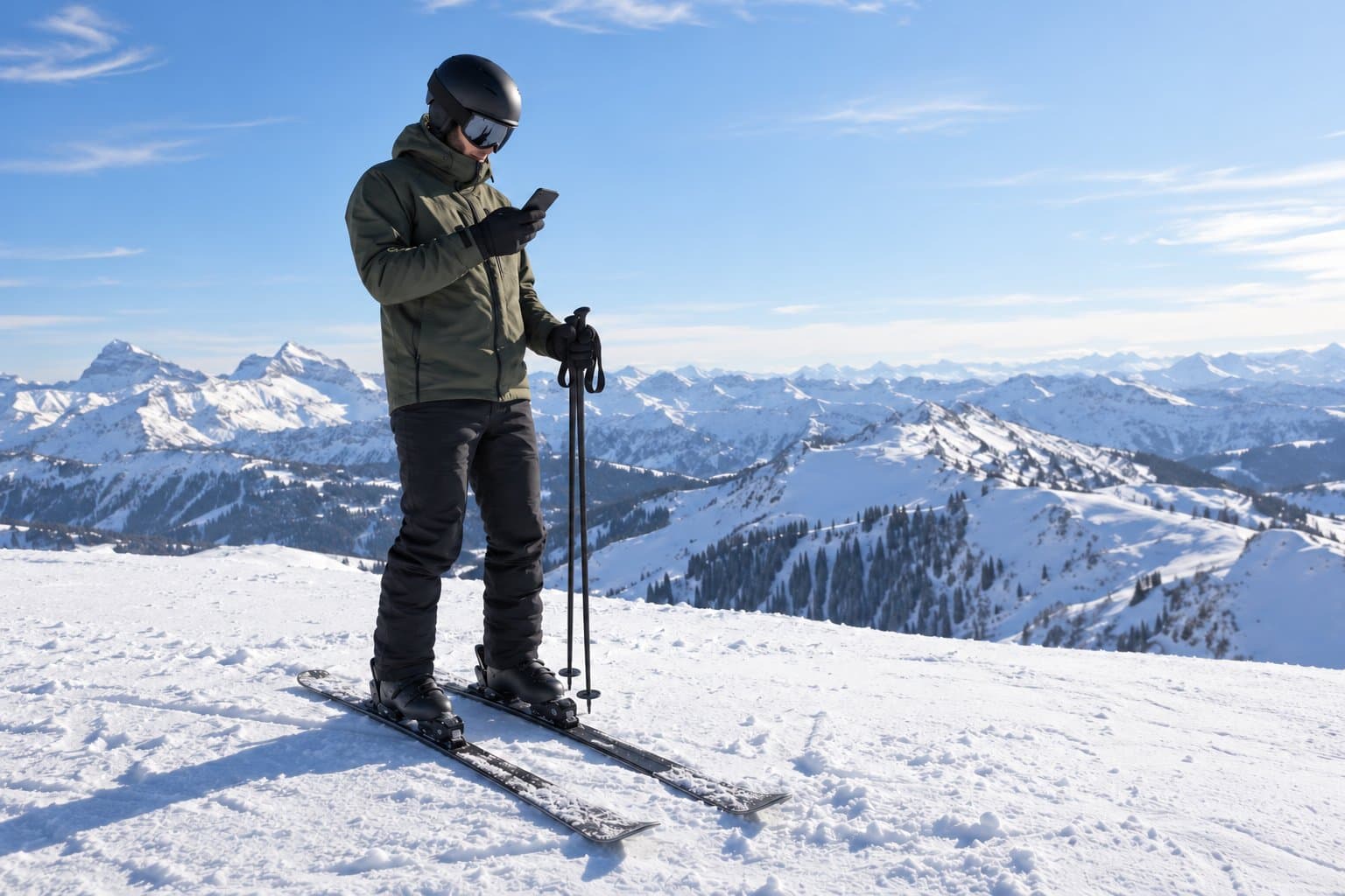 A skier at the top of a run checks a phone, with a wide snowy mountain view behind.